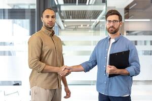 Two businessmen shake hands in an office setting, symbolizing a deal or agreement. The men are well-dressed and appear confident, set against a modern office environment. photo
