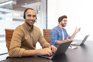 Two diverse customer service representatives wearing headsets are working on laptops in a modern office environment. photo