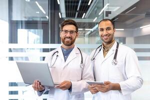 Two smiling doctors pose in a modern office setting, one holding a laptop and the other a tablet. photo