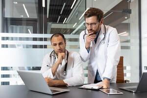 Two doctors, wearing white coats and stethoscopes, look at a laptop screen in a modern office setting, deep in thought. photo