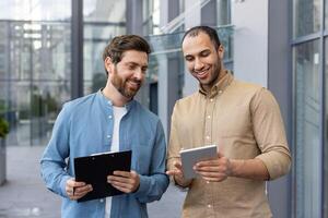 Two diverse businessmen review a tablet, possibly discussing a project or presenting ideas, smiling in a modern outdoor setting. photo