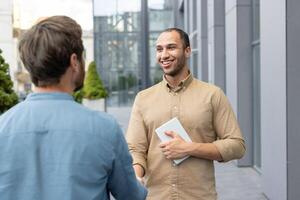 Two businessmen shaking hands in front of a modern building. One man holds a tablet and smiles. photo