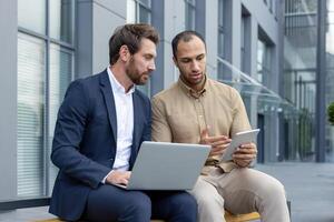 Two diverse businessmen in suits discuss business on a bench outside a modern office building, using laptop and tablet. photo