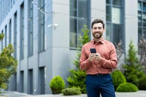 A smiling man holding a phone in front of a modern building. He is outdoors, with greenery. photo