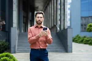 Serious man in salmon shirt and blue pants standing outdoors using mobile phone. He is in front of a building with steps and greenery. photo