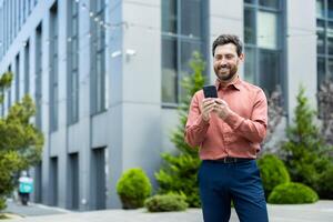 A happy businessman uses his mobile phone outside an office building, smiling as he reads a message or browses the internet. photo