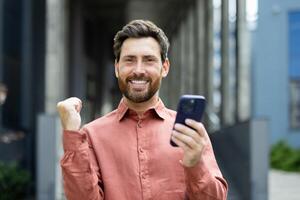 A man with a beard wearing a button-down shirt is holding a smartphone and celebrating success, outdoors with an urban background. photo