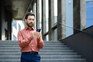 Serious thinking businessman with phone in hands reads online messages and browses internet pages. Man uses smartphone outside office building, walking through city. photo