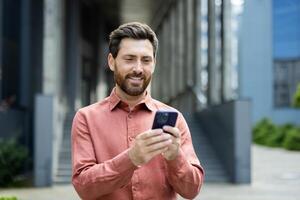 A bearded man smiles while looking at his phone outside a modern building. He is wearing a button-down shirt. photo