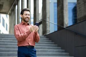 A man in smart casual clothing stands on concrete steps outside a modern building, focused on his mobile phone, smiling at the screen. photo