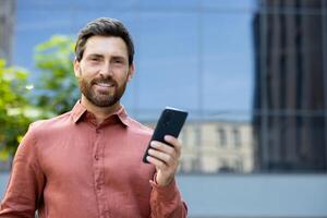 A smiling man stands outdoors, holding a smartphone in front of a reflective building, embodying connection and modern lifestyle. photo