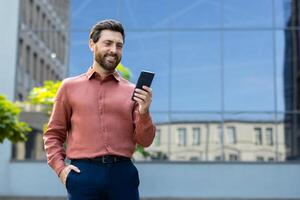 A smiling businessman in a casual outfit looks at his phone in front of a modern building. The man appears to be outside. photo