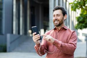 Cheerful man with earphones using a smartphone while standing in the city. The background shows a modern building on a sunny day. photo