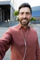 Phone camera view, vertical shot, man in wired headphones looking into smartphone camera, smiling and talking to colleagues and partners remotely, walking outside office building in city photo