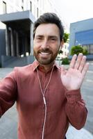 Smiling man with a beard waves while conferencing with earbuds in front of a modern building on a sunny day. photo