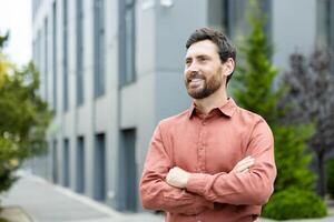 A confident, smiling man with crossed arms stands outside an office building, possibly reflecting on his success and future ventures photo