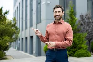 Cheerful man in a terracotta shirt standing outdoors pointing both hands to the left, in front of a modern building and greenery. photo
