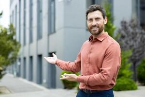 A smiling man in shirt stands in front of a modern building with outstretched hands, inviting viewers in photo