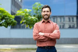 Portrait of successful businessman in shirt, man smiling and looking at camera with crossed arms outside office building. Satisfied with results of achievement, company employee. photo