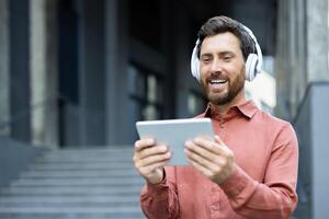Close-up of a joyful man watching online while walking outside an office building. Businessman wearing headphones and holding a tablet computer, smiling happily. photo