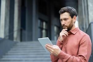 A contemplative man with a beard reviews data on a tablet device in front of a modern, gray building, looking thoughtful and engaged. photo