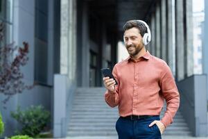 Man with beard enjoys music and mobile apps on his smartphone while walking outside, wearing headphones in a modern urban environment. photo