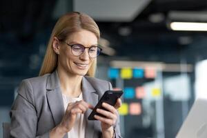 A smiling businesswoman wearing glasses, using a smartphone, likely in a modern office setting during a workday. photo