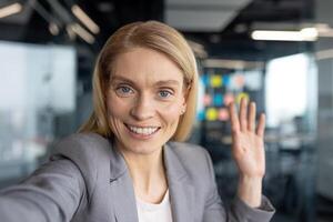 A smiling blonde businesswoman waves hello during a call, with a professional office setting in the background. photo