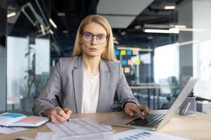 A professional businesswoman in a modern office setting, working on a laptop and taking notes, with a serious expression. photo