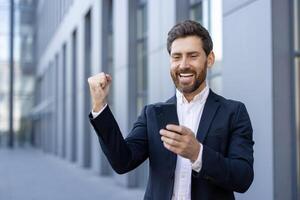 A successful businessman celebrates a win, looking at his phone, with a raised fist and a joyful expression near a modern building. photo