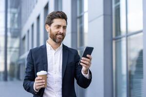 Successful businessman walking through the city with phone in hand, smiling happily. Man in business suit browsing social media and writing text message, with cup of hot drink in hand. photo