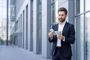 A businessman in a suit counts money in front of a modern building, highlighting wealth and financial success. The man appears thoughtful as he handles the cash. photo