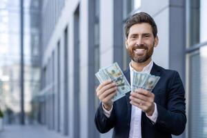 A successful businessman holding cash and smiling confidently in front of an office building, a symbol of wealth. photo