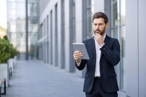 A focused businessman in a suit uses a tablet outside a modern office building, deep in thought. photo