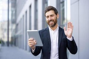 A businessman greets someone on a call, holding a tablet. He smiles while wearing a suit outside a modern building. photo