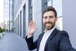 A smiling man waves at the camera while wearing a suit and earpieces, outside a modern building. It's a self-portrait. photo