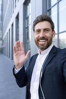 A smiling businessman in a suit waves towards the camera while wearing earbuds outdoors. The background shows an office building. photo