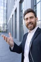 A smiling businessman is on a call in front of a modern building, wearing a suit and using earphones, presenting with his hand photo