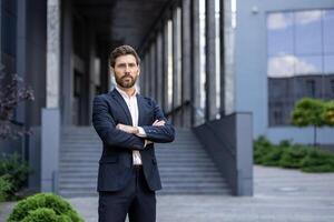 A confident businessman stands outside a modern building, arms crossed, radiating professionalism. He wears a suit, ready for business. photo