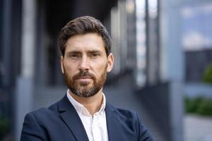 A handsome, bearded man in a suit poses confidently, looking directly at the camera with a composed expression. The urban backdrop hints at a professional setting. photo