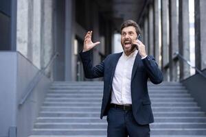A frustrated businessman in a suit yells into his phone while outside, gesturing wildly. The urban setting adds context to his stressful conversation. photo
