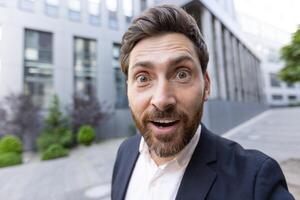 A surprised businessman looks directly at the camera outdoors with his mouth open in shock, in front of a modern building. photo