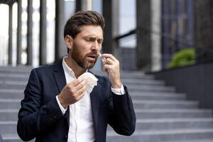 A businessman in a suit outside, looking distressed, blowing his nose with a tissue near a building entrance. photo