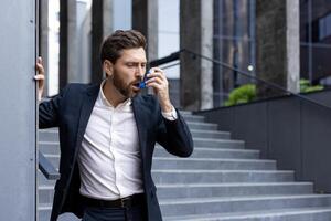 A professional man uses an inhaler outside of a modern office building. He appears to be struggling with a sudden asthma attack or breathing problem while on the stairs. photo
