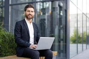 A smiling businessman sits outside, working on a laptop. He wears a suit and is positioned in front of a modern building, suggesting a professional setting. photo
