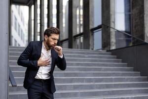 A businessman, clearly unwell, coughs while clutching his chest outdoors. The man is experiencing what seems to be illness or respiratory issues. photo