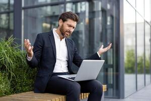 A businessman in a suit, looking confused with a laptop, sitting on a bench near a modern building. He is expressing frustration. photo