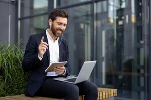 A businessman is working on a laptop outside. He is smiling and holding a notebook while on a call, looking engaged. photo