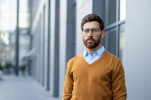 A serious-looking businessman with a well-groomed beard stands in front of a modern building. He is wearing glasses and casual business attire. photo