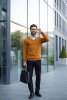 A smiling businessman with glasses adjusts his frames while holding a briefcase outside a modern office building. He wears a smart casual attire. photo